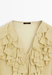 Beige bluse med V-hals og flere kaskader af flæser foran. Blød tekstur med synlige, frynset kanter. Sort mærke indeni.