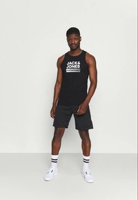 Un homme se tenant debout portant un débardeur noir "Jack & Jones Performance", un short noir, des chaussettes blanches avec des rayures noires et des baskets blanches.