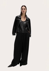 Veste en cuir noire ornée de clous argentés, portée sur un haut noir sans manches, associée à un pantalon noir à jambes larges et des chaussures noires à bouts pointus.