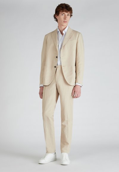 Completo in lino beige con vestibilità sartoriale, dotato di giacca a un petto e pantaloni con frontale piatto, abbinato a sneakers bianche.
