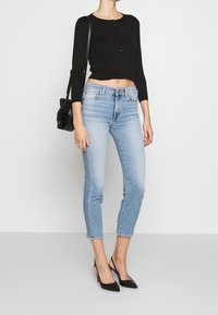 Mujer con un cárdigan negro corto con botones, jeans skinny azul claro, tacones negros de tipo slingback y llevando un bolso de hombro de cuero negro.