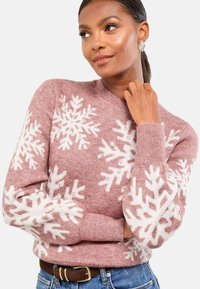 Hellrosa Pullover mit weißen Schneeflockenmuster und Pailletten, langen Ärmeln und Rundhalsausschnitt. Kombiniert mit blauen Jeans und einem braunen Gürtel.