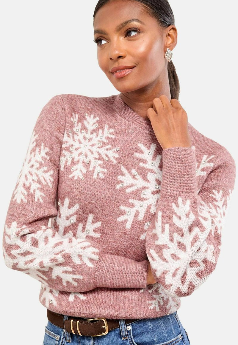 Hellrosa Pullover mit weißen Schneeflockenmuster und Pailletten, langen Ärmeln und Rundhalsausschnitt. Kombiniert mit blauen Jeans und einem braunen Gürtel.