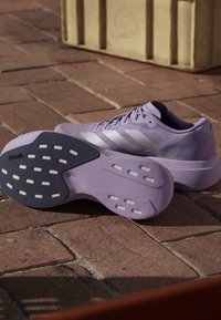 Para jasnofioletowych butów do biegania Adidas z białymi paskami, ukazująca podeszwę z czarno-białym bieżnikiem, umieszczona na ceglanej podłodze.
