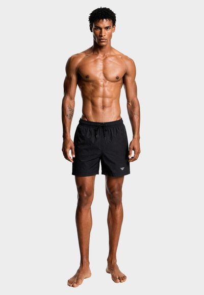 Μυώδης άντρας που στέκεται ξυπόλητος, φορώντας μαύρα μαγιό με κορδόνι και με τατουάζ και στα δύο αντιβράχια, πάνω σε απλό φωτεινό φόντο.