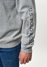 Grå hettegenser med ribbestrikkede mansjetter og nederkant, og marineblå tekst "U.S. POLO ASSN. SINCE 1890" langs venstre arm. Mykt, bomullsblandingsmateriale.