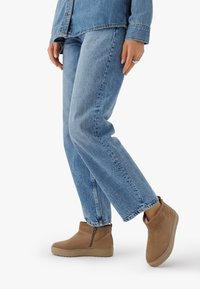 Person, der eine hellblaue Jeans und eine passende Jeansjacke trägt, geht in beigefarbenen Stiefeletten mit seitlichen Reißverschlüssen vor einem weißen Hintergrund.