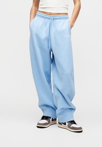 Άτομο που φορά φαρδιά ανοιχτό μπλε παντελόνια φόρμας με κορδόνι και λευκό crop top, με τα χέρια στις τσέπες, συνδυασμένο με μαύρα και λευκά αθλητικά παπούτσια.