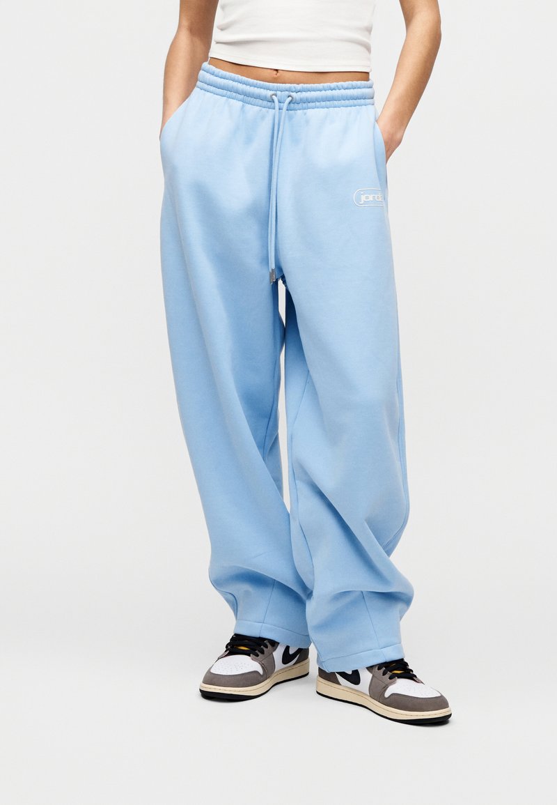 Άτομο που φορά φαρδιά ανοιχτό μπλε παντελόνια φόρμας με κορδόνι και λευκό crop top, με τα χέρια στις τσέπες, συνδυασμένο με μαύρα και λευκά αθλητικά παπούτσια.