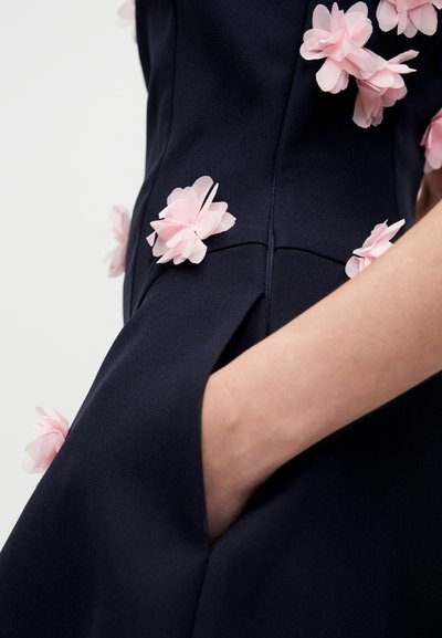 Abito blu navy in tessuto testurizzato, con applicazioni floreali rosa pastello. Include una tasca laterale e una vestibilità sartoriale.