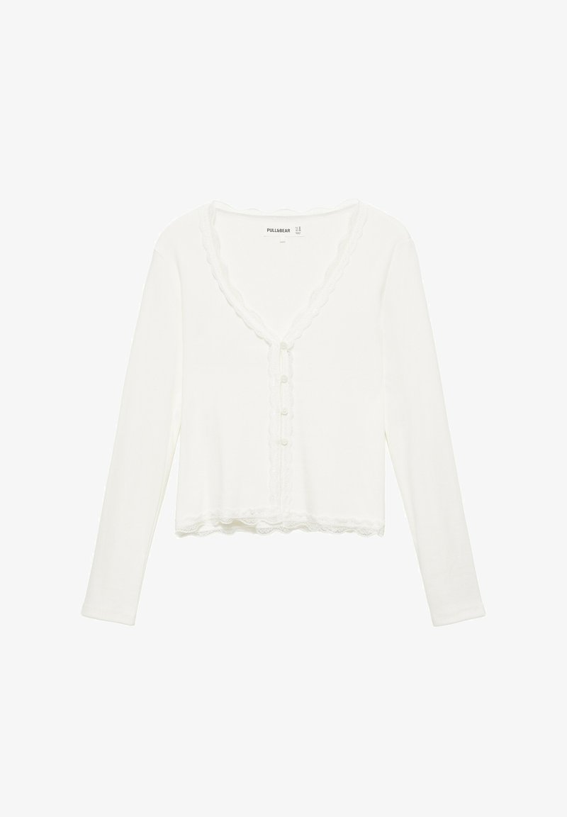 Cardigan court blanc à manches longues avec bords festonnés, boutons sur le devant et une bordure texturée le long de l'encolure et de l'ourlet.