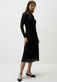 Schwarzes Spitzenkleid mit langen Ärmeln, strukturiertem Muster, leicht ausgestellter Silhouette und einem Saum, der bis zur Mitte der Wade reicht. Getragen mit schwarzen Ballerinas.