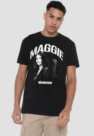 Junger Mann trägt ein schwarzes T-Shirt mit einer Grafik von Maggie aus The Walking Dead, die Pfeile hält, kombiniert mit beigen Cargoshorts.