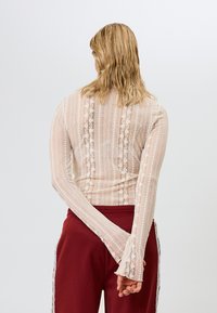 Lys beige langermet topp med et teksturert blondemønster med vertikale striper. Den har tettsittende ermer og en avrundet bakseam.