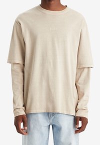 Mężczyzna odziany w beżową, warstwową koszulę z długim i krótkim rękawem marki Levi's, w połączeniu z jasnoniebieskimi dżinsami, stojący na białym tle.