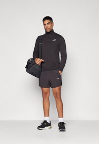Pull-over en polaire noir à demi-zip, shorts noirs et chaussures de sport noires. Comprend un sac de sport noir avec des accents en maille, avec le logo en évidence.