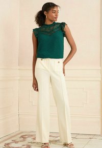 Top senza maniche verde con texture, bordi in pizzo arricciati, abbinato a pantaloni bianchi a gamba larga con due tasche anteriori e bottoni dorati.