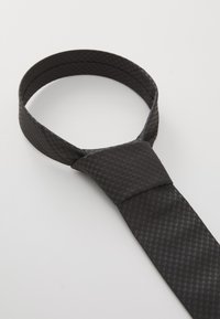 Cravate noire texturée nouée en un nœud Windsor, posée à plat sur une surface blanche.