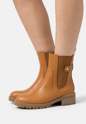 Beige nilkkurit, joissa on joustavat sivupaneelit, sileä nahkapinta ja puumainen blokkikorko. Sivussa on nappiyksityiskohta.