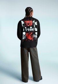 Tricou lung negru cu mâneci, cu imprimeu grafic roșu și alb pe spate, asortat cu pantaloni largi de culoare închisă.