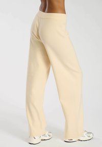 Pantalones anchos color crema con una cinturilla acanalada, hechos de una tela suave, con un ajuste holgado y una textura lisa. Llevados con zapatillas deportivas.