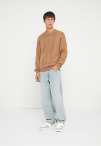 Pull en tricot marron avec un motif de losanges texturé, porté avec un jean baggy bleu clair et des baskets blanches avec des accents beiges.