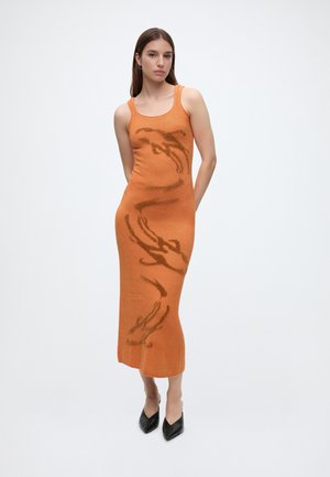 Femme portant une robe midi orange sans manches avec des motifs bruns abstraits, debout les mains dans le dos et des chaussures noires pointues.
