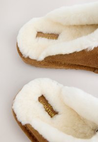 Brązowe zamszowe kapcie z miękką białą sztuczną futrzaną wyściółką i wkładką. Logo marki "SIGNATUR" jest widoczne na złoto po wewnętrznej stronie.