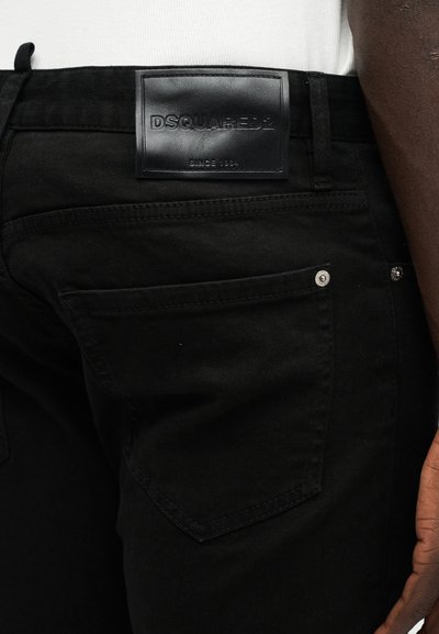 Jean en denim noir porté avec un haut blanc, montrant la poche arrière et le patch en cuir noir de la marque DSQUARED2 sur la ceinture.