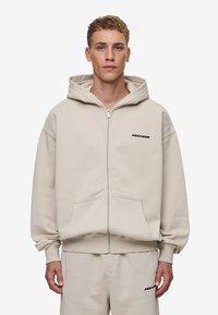 Beige zip-up hættetrøje med en stor hætte, forlommer og sort "PEGADOR" logo. Lavet af et blødt, tekstureret stof med en afslappet pasform.