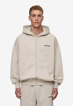 Beige ritsjack met een grote capuchon, voorzakken en een zwart "PEGADOR" logo. Gemaakt van een zachte, gestructureerde stof met een losse pasvorm.