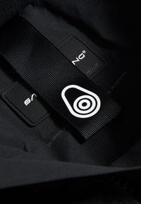 Sort stofmærke med et hvidt logo, syet label der angiver størrelse medium, og tekstdetaljer på en struktureret baggrund.