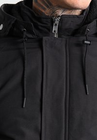 Homme portant une veste noire zippée avec des cordons de serrage et un tatouage visible au niveau du cou.