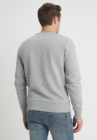 Uomo che indossa una felpa grigia chiara semplice e jeans blu, in piedi rivolto di spalle contro uno sfondo chiaro e uniforme.