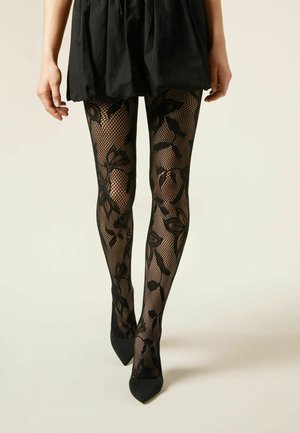 Benen met zwart visnetpanty met bloemenpatroon en zwarte schoenen met puntige neus onder een korte zwarte rok op een lichte achtergrond.