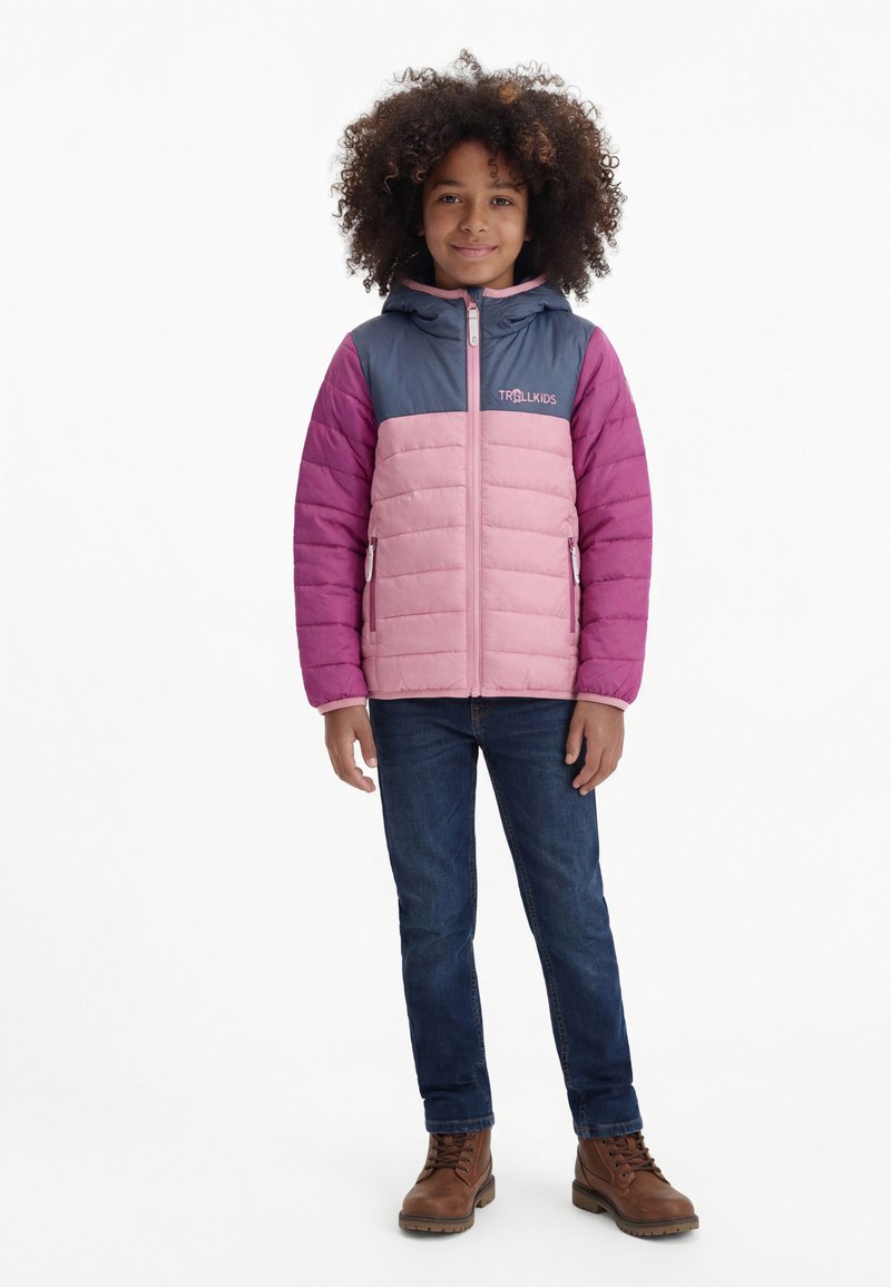 Kind mit lockigem Haar, das eine rosa-lila Kapuzenjacke, blaue Jeans und braune Stiefel trägt, steht vor weißem Hintergrund und lächelt.