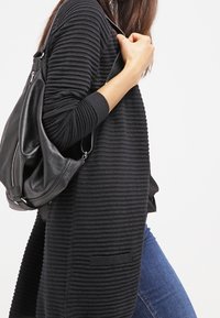 Schwarze gerippte Strickjacke mit langen Ärmeln, vorderen Taschen und offener Vorderseite, kombiniert mit einer schwarzen Schultertasche und blauen Jeans.