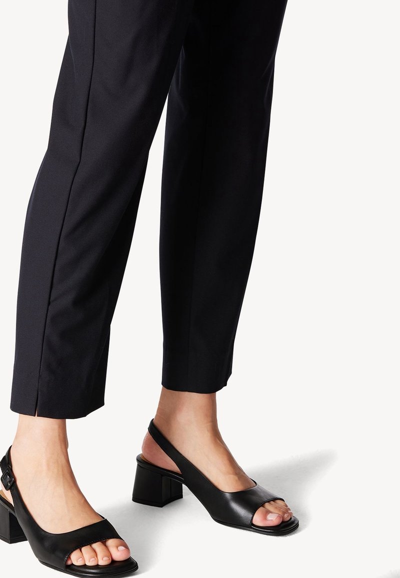 Jambes portant un pantalon noir raccourci et des chaussures à talons carrés ouvertes à l'avant avec bride arrière, sur fond blanc.