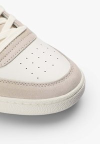 Primer plano de la punta de una zapatilla blanca con ribete de ante beige, parte superior de cuero perforado, cordones blancos y suela de goma texturizada sobre fondo blanco.