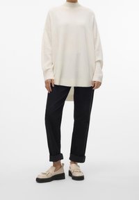 Personne portant un pull crème oversize, un pantalon noir retroussé et des mocassins beige à franges, debout devant un fond blanc uni.