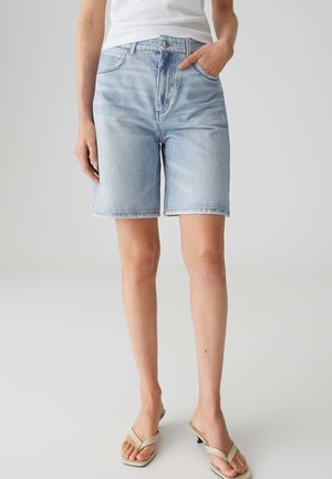 Persona che indossa pantaloncini di jeans azzurri fino al ginocchio, top bianco senza maniche e sandali beige con tacco, in piedi con una mano in tasca.