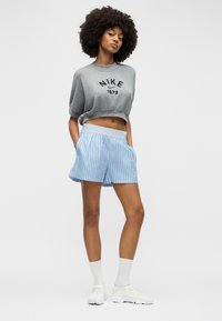 Νεαρή γυναίκα με αφρο μαλλιά που φοράει γκρι κοντό φούτερ Nike, ανοιχτό μπλε ριγέ σορτς, λευκές κάλτσες και λευκά αθλητικά παπούτσια, στέκεται με τα χέρια στις τσέπες.