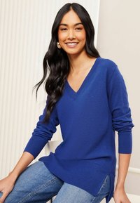 Maglione blu a scollo a V in tessuto a maglia con polsini e orlo a costine, caratterizzato da spacchi laterali. Indossato con jeans blu, seduto contro uno sfondo chiaro.