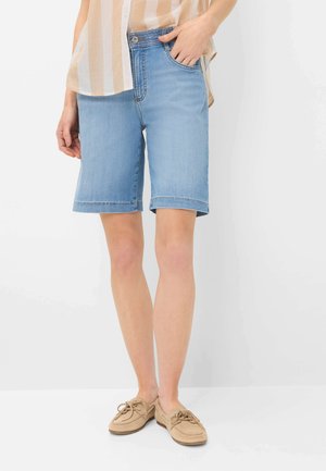 Person iført lyseblå denim Bermuda shorts, beige stribet skjorte og brune mokkasiner, stående med den ene hånd i lommen.