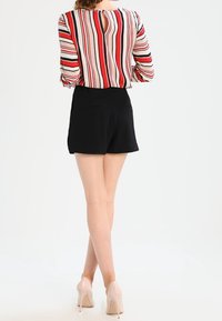 Blouse rayée avec des lignes verticales rouges, blanches et noires, dotée d'une ouverture en trou de serrure à l'arrière, assortie à un short noir et des talons nus.