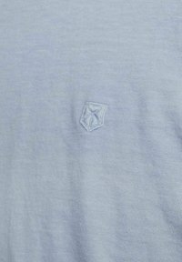 Light blue cotton T-shirt featuring a small embroidered shield logo on the left chest area. The fabric has a soft, smooth texture.