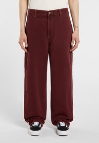 Pantalones de pierna ancha burdeos con cierre de botón, doble bolsillo lateral y costuras amarillas contrastantes, combinados con zapatillas negras y blancas.