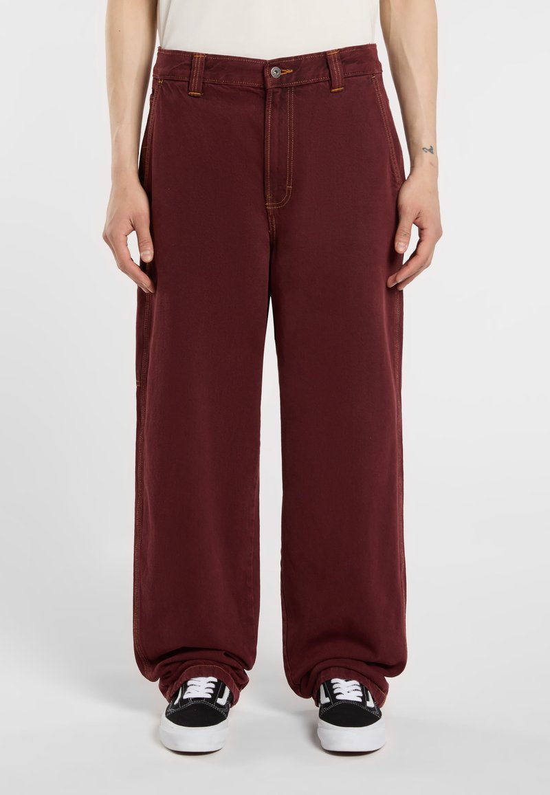 Pantalones de pierna ancha burdeos con cierre de botón, doble bolsillo lateral y costuras amarillas contrastantes, combinados con zapatillas negras y blancas.