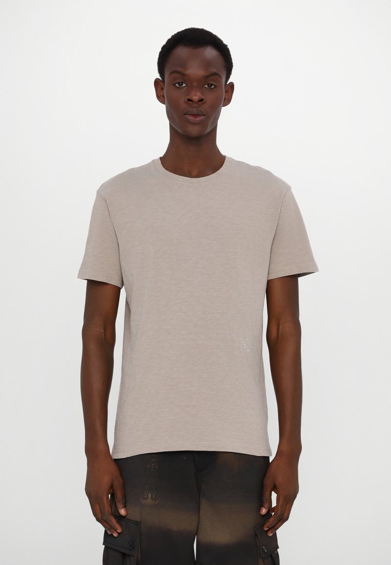 Beige, kortärmad T-shirt med rund halsringning, tillverkad av mjukt tyg. Innehåller en liten grafisk detalj nära fållen.
