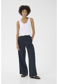 Weißes Tanktop, marineblaue weite Hose und schwarze Sandalen. Glatter Stoff mit lockerer Passform. Das Haar ist lockig, und das Model steht mit einer Hand auf der Hüfte.